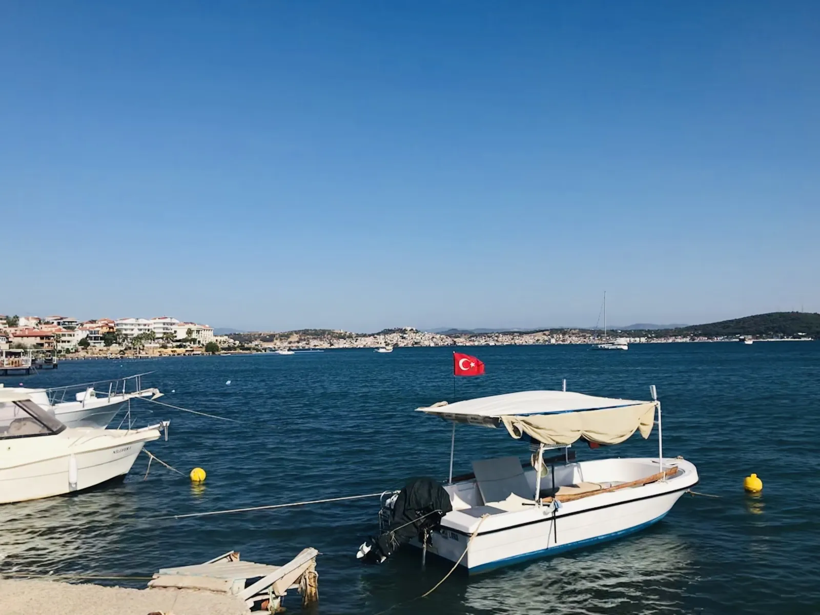 Cunda Adası kıyısında bağlı küçük tekneler, mavi deniz ve karşı kıyıdaki yerleşim